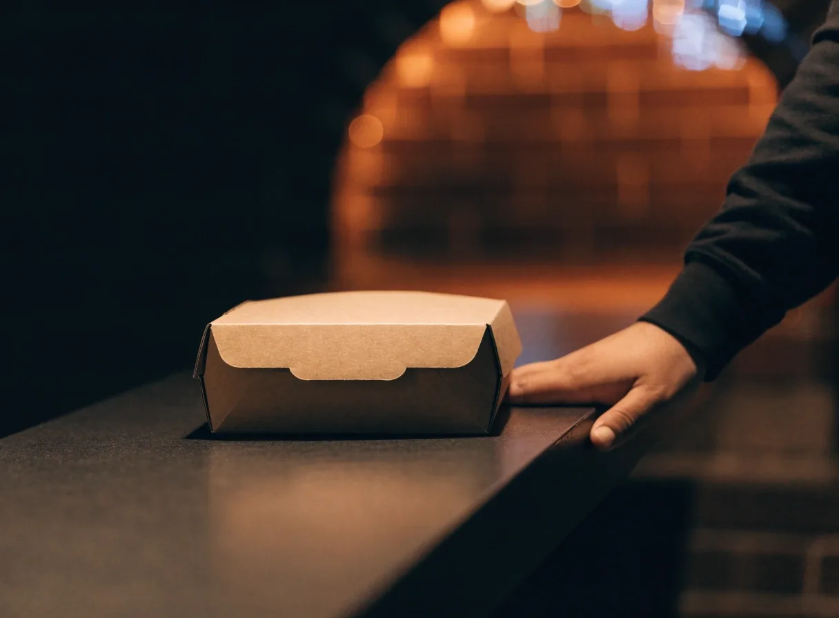 Closed takeout box resting on a dark counter with a hand at the edge